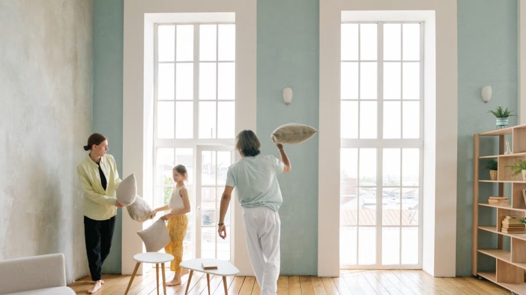 Playful family pillow fight in a bright modern living room with large windows.