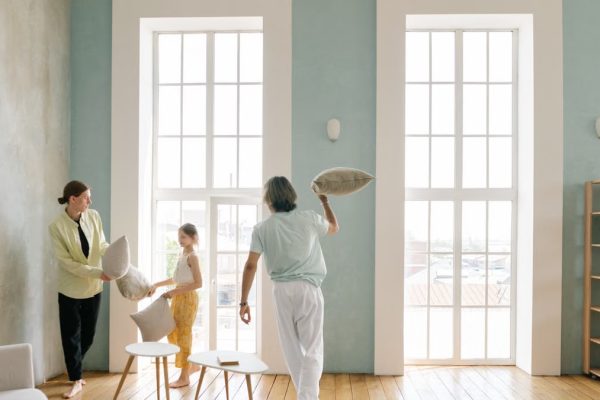 Playful family pillow fight in a bright modern living room with large windows.